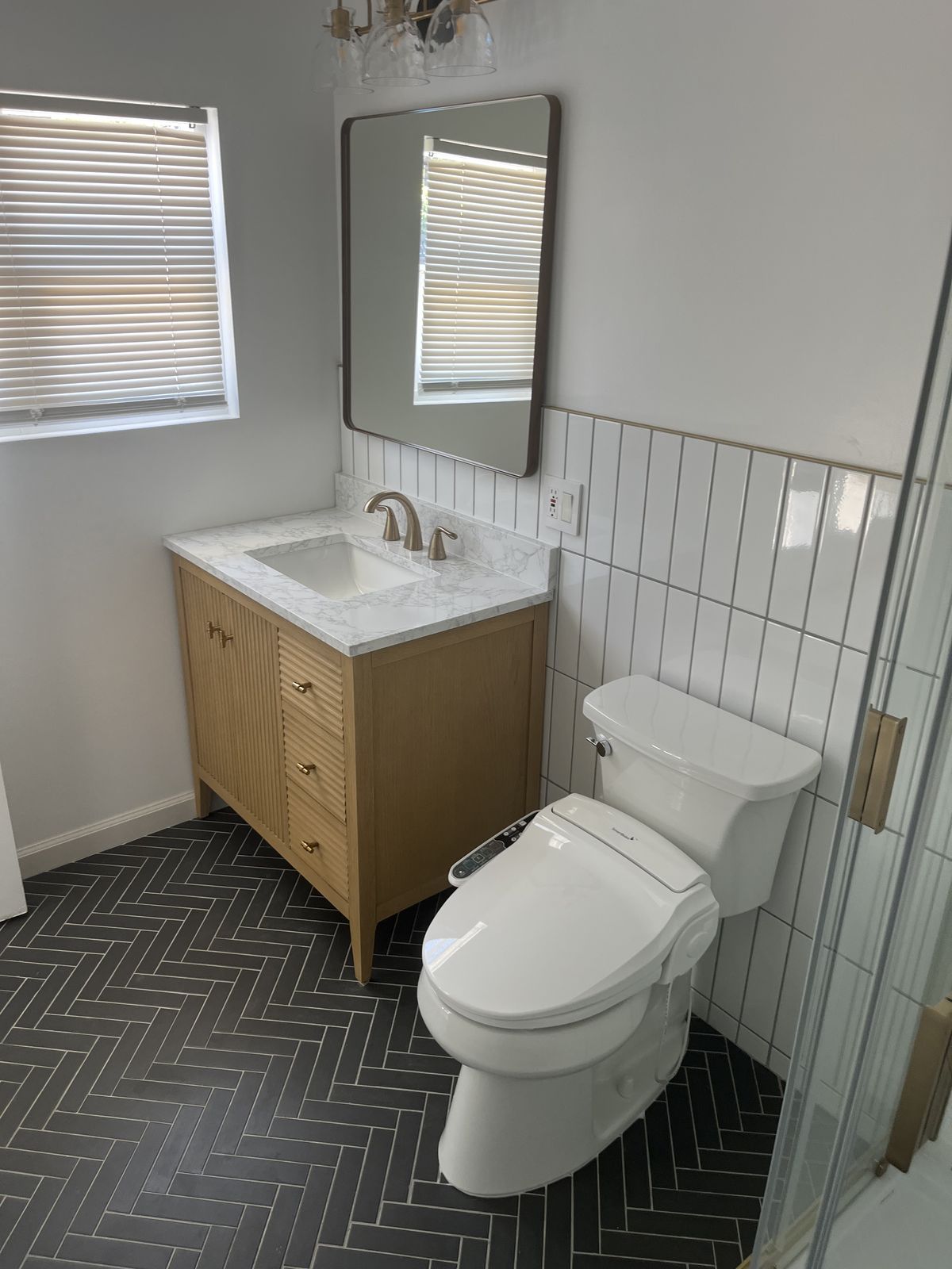 Completed bathroom vanity installation in San Mateo showing light wood cabinet with marble countertop, herringbone tile floor, and white subway tile wainscoting